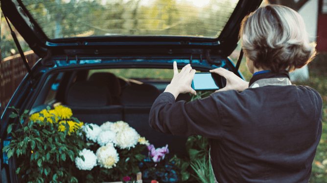 Woman using her mobile device to take a picture of flowers in the trunk of her car.
